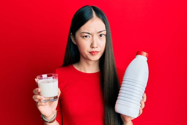Jovem chinesa bebendo um copo de leite fresco cético e nervoso, franzindo a testa por causa do problema. pessoa negativa.  - Foto, Imagem