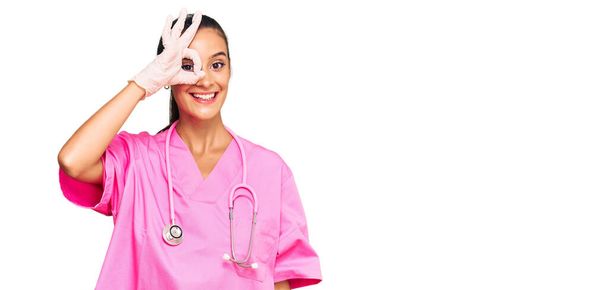 Молодая латиноамериканка в докторской форме и стетоскопе улыбается счастливо делая хорошо знак с рукой на глазу глядя сквозь пальцы  - Фото, изображение