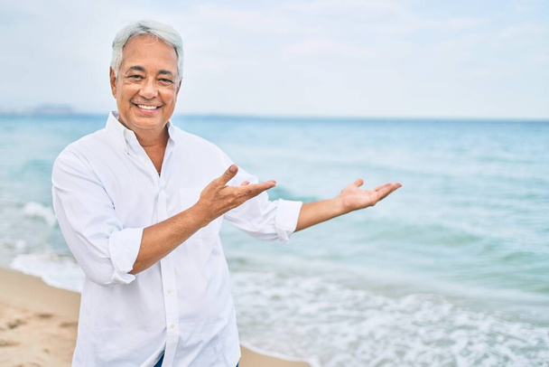 Handosme Ισπανόφωνος άνδρας με γκρίζα μαλλιά χαμογελώντας χαρούμενος στην παραλία, απολαμβάνοντας τις διακοπές δείχνοντας χώρο αντίγραφο - Φωτογραφία, εικόνα