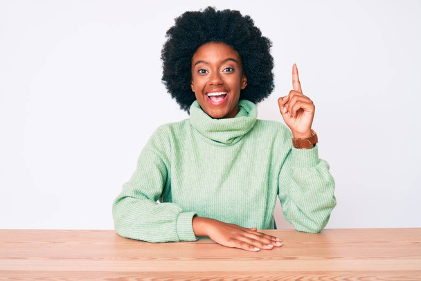 Mujer afroamericana joven con suéter casual de invierno apuntando con el dedo hacia arriba con una idea exitosa. exitado y feliz. número uno.  - Foto, imagen