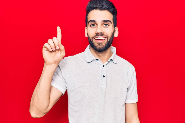 Junger schöner Mann mit Bart trägt lässiges Polo und zeigt mit Finger Nummer eins nach oben, während er selbstbewusst und glücklich lächelt.  - Foto, Bild