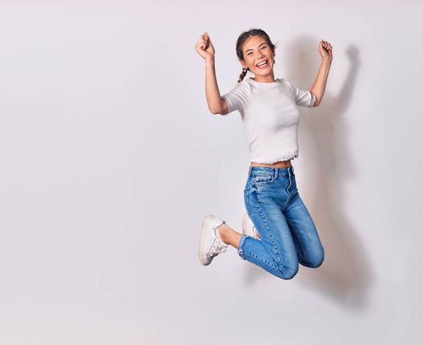 Young beautiful woman with braids wearing casual clothes smiling happy. Jumping with smile on face doing winner sign with fists up over isolated white background - Photo, Image