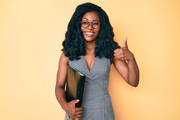 Bella donna africana in possesso di cartella aziendale sorridente felice e positivo, pollice in su facendo segno eccellente e approvazione  - Foto, immagini