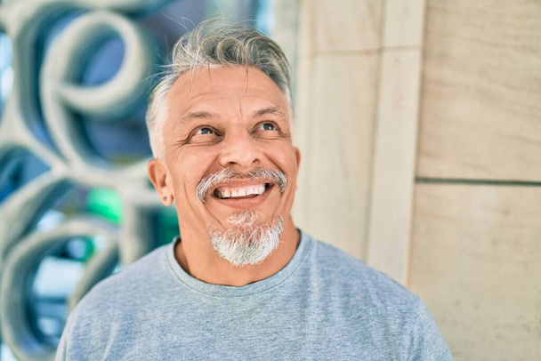 Hombre de pelo gris hispano de mediana edad sonriendo feliz de pie en la ciudad. - Foto, imagen