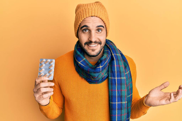 Joven hombre hispano usando suéter de lana y bufanda de invierno sosteniendo píldoras celebrando el logro con sonrisa feliz y expresión ganadora con la mano levantada  - Foto, imagen