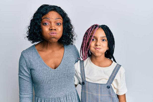 Beautiful african american mother and daughter wearing casual clothes and hugging puffing cheeks with funny face. mouth inflated with air, crazy expression.  - Foto, imagen
