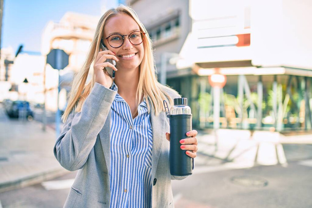 Jeune femme d'affaires blonde parlant sur le smartphone et tenant une bouteille d'eau à la ville. - Photo, image