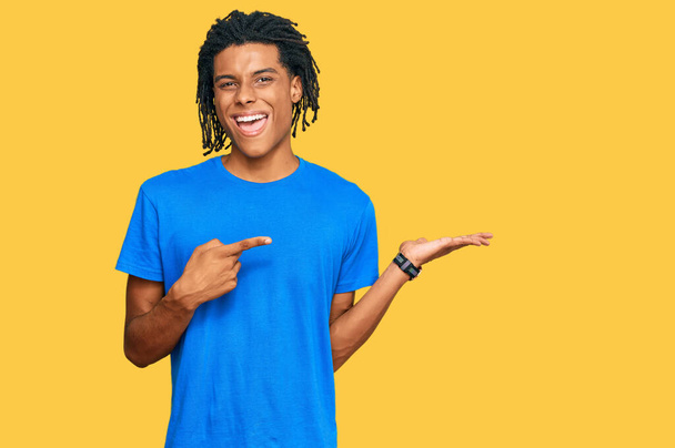 Junger afrikanisch-amerikanischer Mann in lässiger Kleidung staunt und lächelt in die Kamera, während er mit der Hand präsentiert und mit dem Finger zeigt.  - Foto, Bild