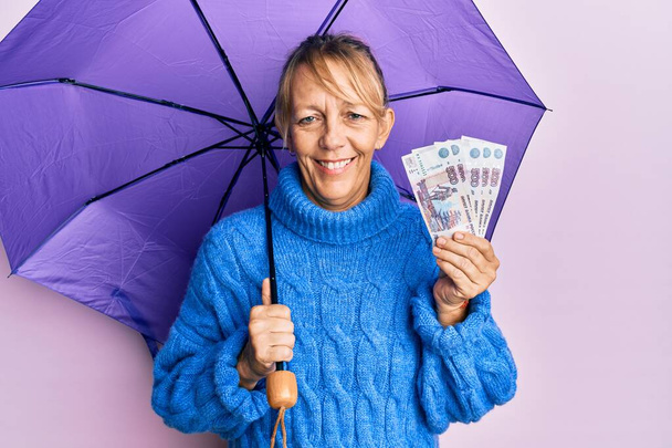 Mujer rubia de mediana edad sosteniendo paraguas y billetes de rublo rusos sonriendo con una sonrisa feliz y fresca en la cara. mostrando dientes.  - Foto, imagen