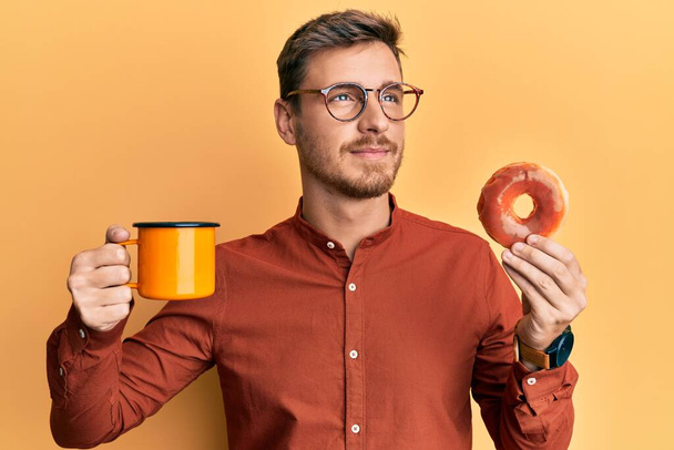 Schöner kaukasischer Mann, der Donut isst und Kaffee trinkt, lächelnd zur Seite schaut und nachdenklich wegstarrt.  - Foto, Bild
