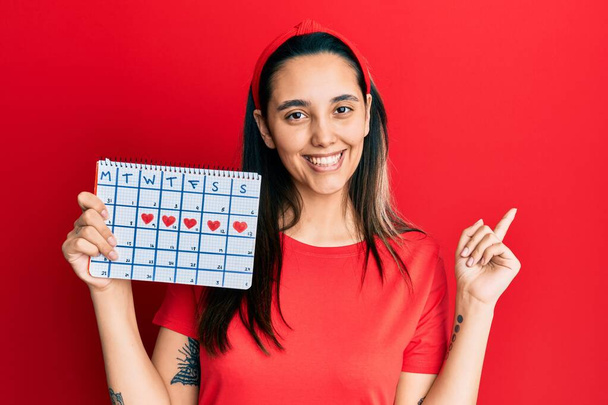 Jeune femme hispanique tenant calendrier cardiaque souriant heureux pointant avec la main et le doigt sur le côté  - Photo, image