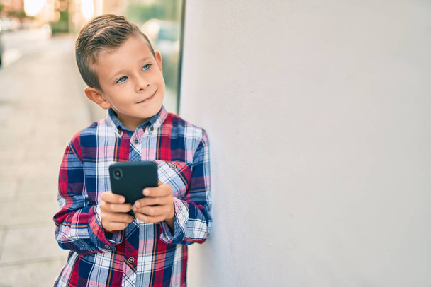 Beyaz çocuk şehirde akıllı telefon kullanarak mutlu bir şekilde gülümsüyor.. - Fotoğraf, Görsel