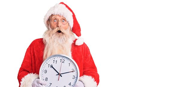 Старик с седыми волосами и длинной бородой в костюме Санта-Клауса держит часы испуганными и изумленными с открытым ртом для удивления, неверного лица  - Фото, изображение