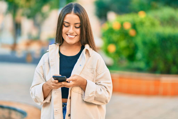 Młoda Latynoska dziewczyna uśmiechnięta szczęśliwy za pomocą smartfona w parku. - Zdjęcie, obraz