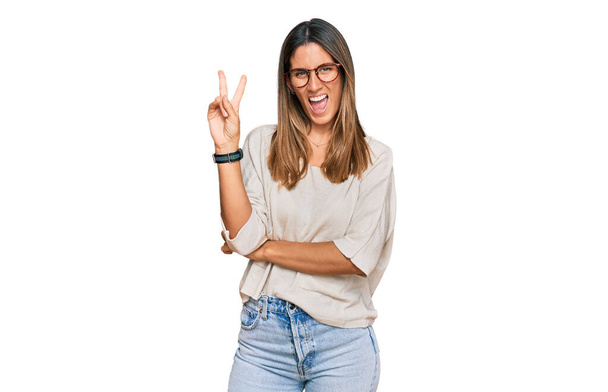 Young woman wearing casual clothes and glasses smiling with happy face winking at the camera doing victory sign. number two.  - Photo, Image