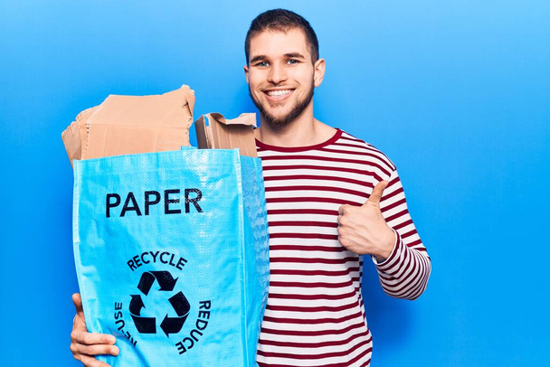 Jovem homem bonito reciclando papel sorrindo feliz e positivo, polegar para cima fazendo excelente e sinal de aprovação  - Foto, Imagem
