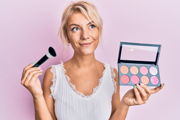 Young blonde girl holding makeup brush and blush smiling looking to the side and staring away thinking.  - Photo, Image