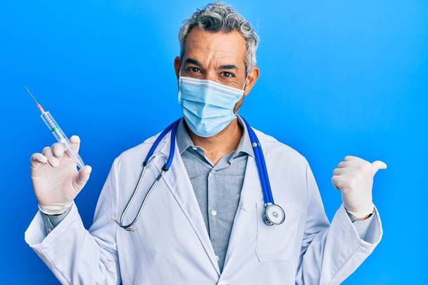 Middle age grey-haired man wearing doctor uniform and medical mask holding syringe pointing thumb up to the side smiling happy with open mouth  - 写真・画像