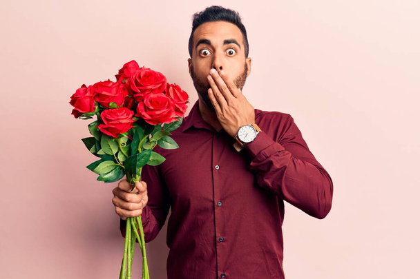 Mladý hispánec držící květiny na ústech rukou, šokovaný a vystrašený z omylu. překvapený výraz  - Fotografie, Obrázek