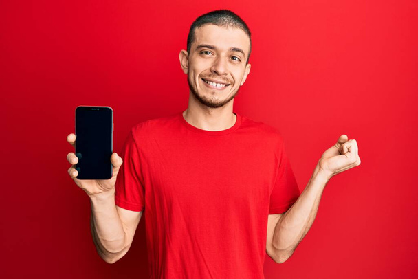 Hispanic young man holding smartphone showing blank screen screaming proud, celebrating victory and success very excited with raised arm  - Φωτογραφία, εικόνα