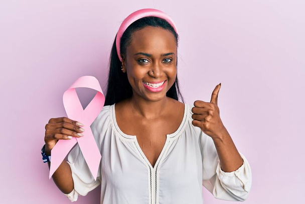 Young african woman holding pink cancer ribbon smiling happy and positive, thumb up doing excellent and approval sign  - Zdjęcie, obraz