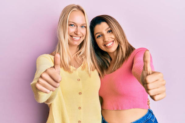Двоє друзів, що стоять разом на рожевому тлі, схвалюють позитивний жест рукою, великі пальці посміхаються і щасливі за успіх. жест переможця
.  - Фото, зображення