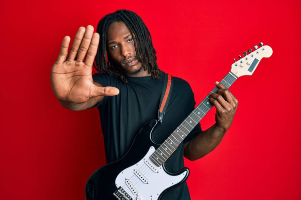 Afro-Amerikaanse man met vlechten spelen elektrische gitaar met open hand doen stopteken met serieuze en zelfverzekerde uitdrukking, verdediging gebaar  - Foto, afbeelding