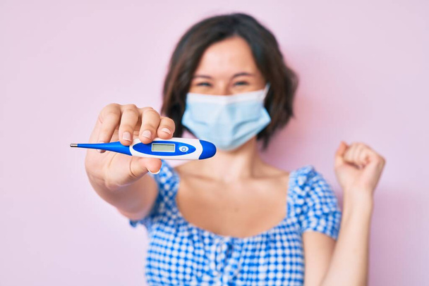 Young beautiful woman wearing medical mask holding holding thermometer screaming proud, celebrating victory and success very excited with raised arms  - Photo, Image