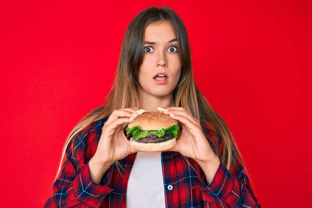 Güzel, beyaz bir kadın şok suratlı, leziz bir klasik burger yiyor, şüpheci ve alaycı görünüyor, ağzı açık kalınca şaşırıyor.  - Fotoğraf, Görsel