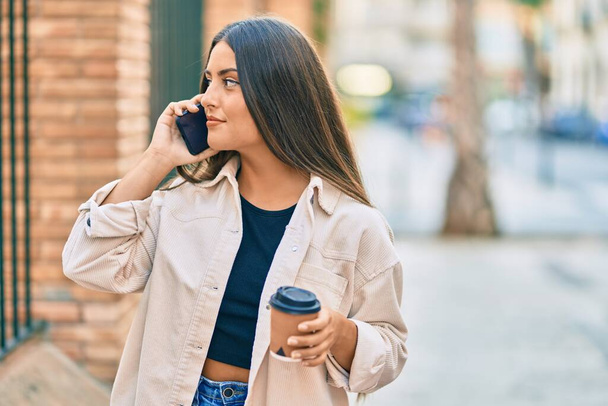 Νεαρή ισπανόφωνη κοπέλα χαμογελά ευτυχισμένη μιλώντας στο smartphone και πίνοντας παίρνει μακριά καφέ στην πόλη. - Φωτογραφία, εικόνα