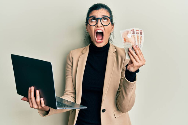 Young hispanic woman wearing business style holding laptop and united kingdom pounds angry and mad screaming frustrated and furious, shouting with anger looking up.  - Foto, Bild