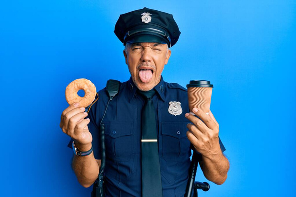 Handsome middle age mature police man eating donut and drinking coffee sticking tongue out happy with funny expression.  - Zdjęcie, obraz