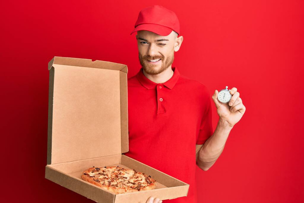 Молодой рыжий мужчина в униформе доставки держит коробку пиццы и часы обратного отсчета подмигивая глядя на камеру с сексуальным выражением лица, веселым и счастливым лицом.  - Фото, изображение