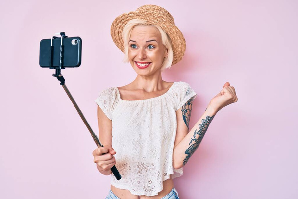 Молодая блондинка с татуировкой делает селфи фото со смартфоном крича гордо, празднуя победу и успех очень взволнован с поднятой рукой  - Фото, изображение
