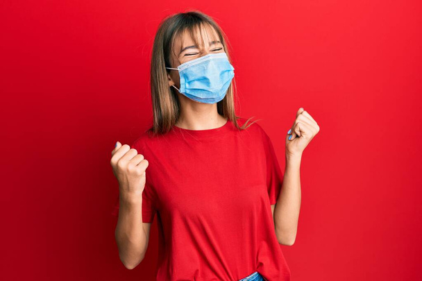 Teenager caucasian girl wearing medical mask very happy and excited doing winner gesture with arms raised, smiling and screaming for success. celebration concept.  - Foto, imagen