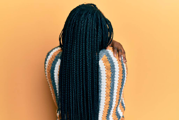 Jeune femme noire avec des tresses portant un pull d'hiver décontracté se serrant dans ses bras heureux et positifs à partir de l'arrière. amour de soi et soins de soi  - Photo, image
