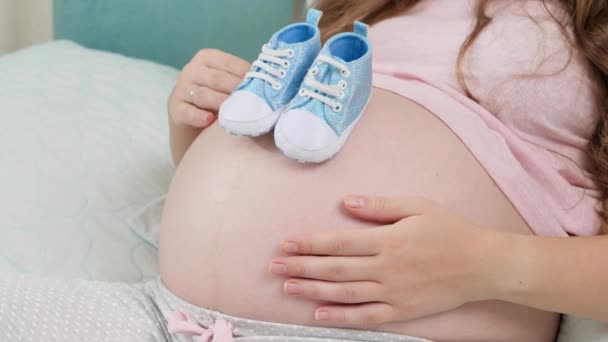 ベビーシャワーの概念。大きな腹に青いベビーブーツを着た赤ん坊を期待している若い妊婦。妊娠の概念,赤ちゃんの準備と期待 - 映像、動画