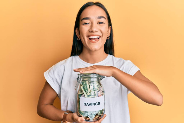 Young asian woman holding jar with savings smiling and laughing hard out loud because funny crazy joke.  - Photo, Image