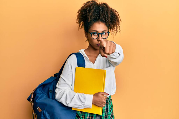 Beautiful african american woman with afro hair wearing school bag and holding books pointing with finger to the camera and to you, confident gesture looking serious  - Fotografie, Obrázek