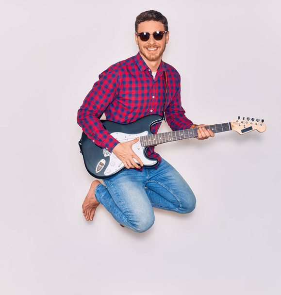 Młody, przystojny, latynoski artysta w okularach słonecznych, uśmiechnięty, szczęśliwy grając na gitarze elektrycznej. Skoki z uśmiechem na twarzy na odizolowanym białym tle - Zdjęcie, obraz