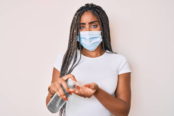 Mujer afroamericana joven con trenzas que usan máscara médica usando desinfectante de manos sin idea y expresión confusa. concepto de duda.  - Foto, Imagen