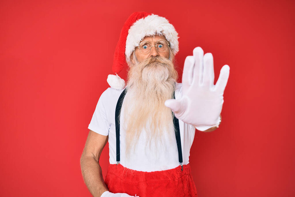 Старик с седыми волосами и длинной бородой в белой футболке и костюме Санта Клауса делает остановку петь с ладонью. предупреждающее выражение с негативным и серьезным жестом на лице.  - Фото, изображение