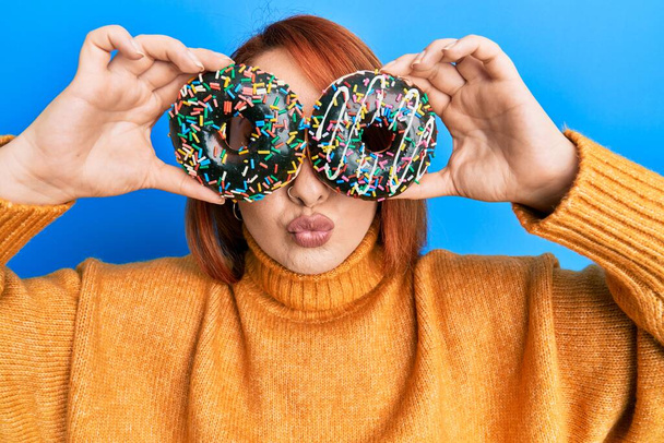 Mulher ruiva bonita segurando saborosos donuts coloridos nos olhos olhando para a câmera soprando um beijo sendo adorável e sexy. expressão do amor.  - Foto, Imagem