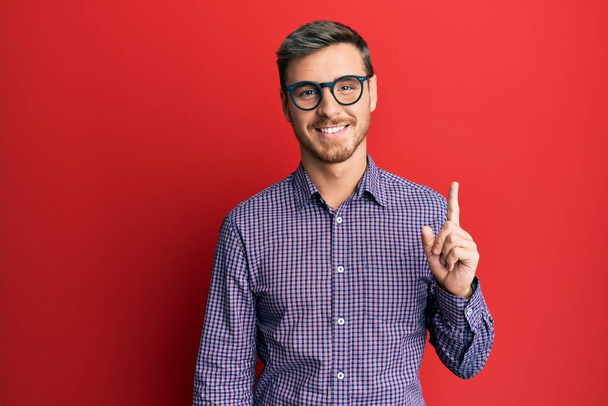 Όμορφος καυκάσιος άντρας που φοράει επιχειρηματικό πουκάμισο και γυαλιά χαμογελώντας με μια ιδέα ή ερώτηση δείχνοντας το δάχτυλο με χαρούμενο πρόσωπο, νούμερο ένα  - Φωτογραφία, εικόνα
