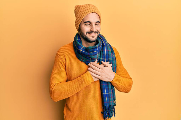 Giovane uomo ispanico che indossa maglione di lana e sciarpa invernale sorridente con le mani sul petto, gli occhi chiusi con gesto grato sul viso. concetto di salute.  - Foto, immagini