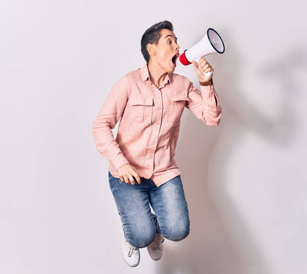 Young beautiful caucasian woman screaming using megaphone. Jumping over isolated white background - Photo, Image