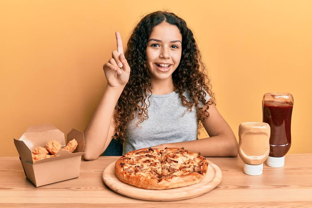 Ragazza ispanica adolescente che mangia pizza e pollo fritto sorridente con un'idea o una domanda che punta il dito verso l'alto con la faccia felice, numero uno  - Foto, immagini