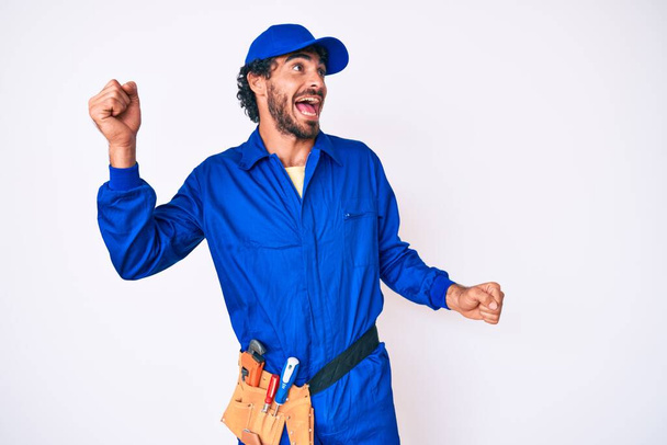 Handsome young man with curly hair and bear weaing handyman uniform dancing happy and cheerful, smiling moving casual and confident listening to music  - Photo, Image