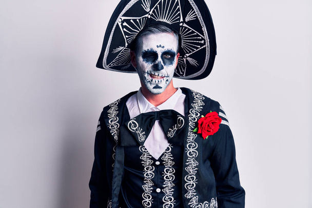 Junger Mann trägt mexikanisches Totenkostüm über weißem Augenzwinkern und blickt mit sexy Gesichtsausdruck, fröhlichem und glücklichem Gesicht in die Kamera.  - Foto, Bild