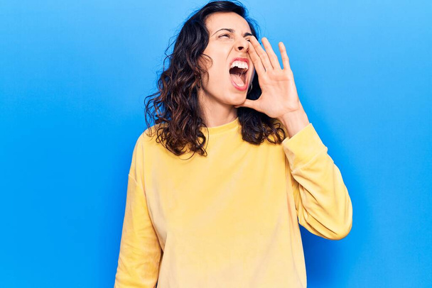 Junge schöne hispanische Frau in lässiger Kleidung schreit und schreit laut mit der Hand auf dem Mund zur Seite. Kommunikationskonzept.  - Foto, Bild